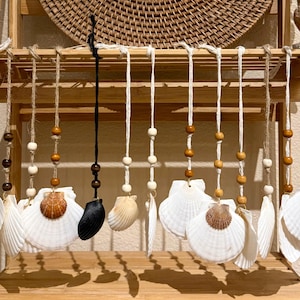 May include: A decorative hanging mobile featuring white seashells, wooden beads, and a black shell. The shells are suspended from twine and arranged on a wooden ladder-style shelf. A woven basket is visible in the background.