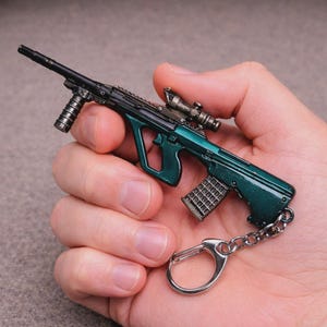 May include: A miniature dark green rifle keyring featuring a detailed barrel, a vertical foregrip, a mounted scope, and a textured magazine. The metal firearm replica is attached to a silver-toned split ring by a short metal chain link.