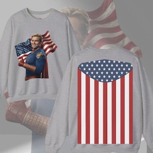 May include: A gray sweatshirt featuring a graphic of a superhero holding an American flag on the front. The back of the sweatshirt displays a stylized American flag design in red, white, and blue. The sweatshirt is a crew neck style.