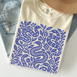 May include: A cream-colored t-shirt featuring a blue graphic print of multiple snakes intertwined with leaves, berries, and small flowers. The shirt has a Comfort Colors label at the inner neckline. Also included are light blue denim jeans and a tan hat.