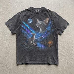 May include: A charcoal grey short-sleeve t-shirt featuring a graphic print of two cats in a forest landscape at night. The scene includes a full moon, blue and purple aurora borealis, a calm river, and a small campfire near the pine trees.