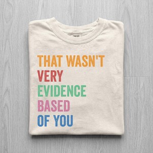 May include: Cream-colored t-shirt with the phrase "THAT WASN'T VERY EVIDENCE BASED OF YOU" in orange, red, green, pink, and blue block letters. The shirt is folded neatly on a light gray wooden surface.