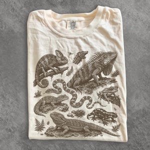 May include: A cream-coloured t-shirt featuring a detailed brown ink illustration of various reptiles and amphibians, including a chameleon, an iguana, a snake, a gecko, a bearded dragon, a frog, and a salamander, with a Comfort Colors brand label.