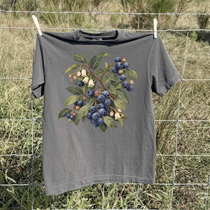 Op de afbeelding: Een antracietgrijs T-shirt met korte mouwen en een centrale grafische illustratie van een bosbessentak. Het ontwerp toont trossen donkerblauwe bessen, groene bladeren en kleine crèmekleurige klokvormige bloemen aan bruine steeltjes.
