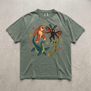 Può includere: Una t-shirt a maniche corte color verde salvia con una stampa grafica raffigurante un Bigfoot marrone, una creatura falena nera, un mostro marino blu e un alieno verde che si tengono per mano in cerchio, circondati da piccole stelle arancioni a quattro punte.