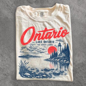 May include: A cream-colored t-shirt featuring a graphic print with the red text Ontario above the blue text Lake Ontario The Great Lakes. The illustration depicts a lighthouse, pine trees, a red sun, clouds, and a rocky shoreline by water.