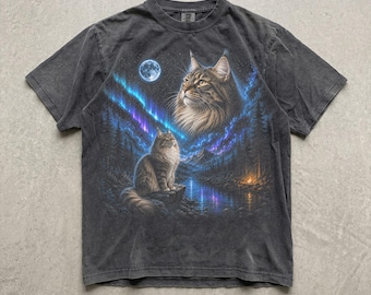 May include: A charcoal gray short-sleeve t-shirt featuring a graphic print of two long-haired cats in a forest landscape. The scene includes a full moon, a vibrant blue and purple aurora borealis, a calm river, and a small campfire in the distance.