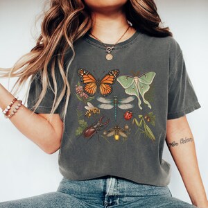 Peut inclure: Un t-shirt gris anthracite à manches courtes avec une impression graphique représentant divers insectes et plantes, dont un papillon monarque, un papillon lune, une libellule, une abeille, une coccinelle, un lucane, une luciole et une mante religieuse.