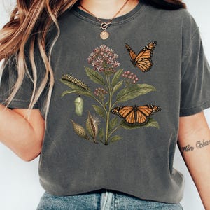 May include: A charcoal gray short-sleeve t-shirt featuring a botanical illustration of a milkweed plant with a monarch caterpillar, a chrysalis, two monarch butterflies, and seed pods. The graphic is centered on the chest of the garment.