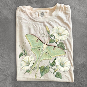 May include: A cream-colored t-shirt featuring a graphic illustration of a pale green luna moth with long tails, surrounded by white morning glory flowers and green leaves. The inner neck label displays the text Comfort Colors.