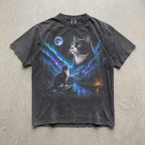 May include: A charcoal gray short-sleeve t-shirt featuring a graphic print of a black and white cat sitting on a rock by a lake, with a larger cat face above, all set against a night sky with a full moon and vibrant blue and purple aurora borealis.