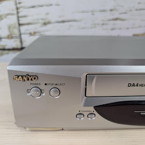 May include: Silver Sanyo VCR with the text "DA4 HEAD Hi-Fi VCR" and the brand name "SANYO" in gold. The VCR has buttons for power, stop/eject, and channel selection. The VCR is on a wooden surface.