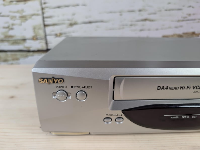 May include: Silver Sanyo VCR with the text "DA4 HEAD Hi-Fi VCR" and the brand name "SANYO" in gold. The VCR has buttons for power, stop/eject, and channel selection. The VCR is on a wooden surface.