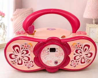 May include: A pink and peach-colored Barbie CD player with a handle, butterfly accent, and floral designs. The player has a digital display and a central flower-shaped control panel. The overall design is bright and playful, suitable for children.
