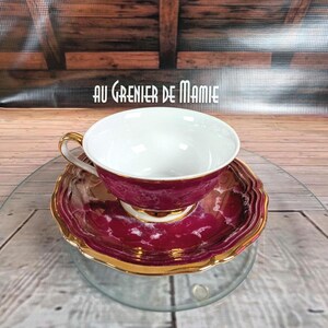 May include: A vintage teacup and saucer set. The teacup and saucer are a deep burgundy colour with a marbled effect and gold trim. The teacup has a white interior and a gold handle. The saucer has a scalloped edge. The text "AU GRENIER DE MAMIE" is visible.