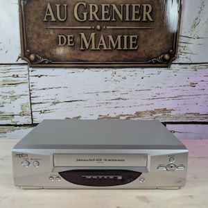 May include: A silver Sanyo VCR with a black and silver front panel. The VCR has a tape slot, control buttons, and the text "DA4 Hi-Fi VCR 19 Micron Head". The background features a wooden surface and a sign that reads "AU GRENIER DE MAMIE".