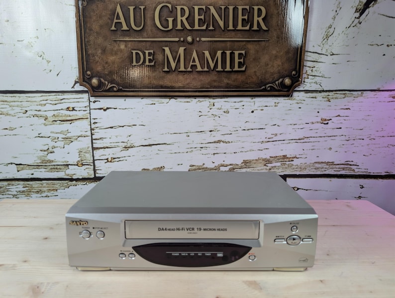 May include: A silver Sanyo VCR with a black and silver front panel. The VCR has a tape slot, control buttons, and the text "DA4 Hi-Fi VCR 19 Micron Head". The background features a wooden surface and a sign that reads "AU GRENIER DE MAMIE".
