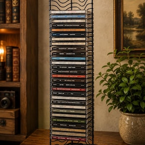 May include: A tall, black metal wire rack designed for vertical storage of compact discs. The structure features a series of horizontal wire shelves held by four vertical supports with rounded finials at the top, holding a stack of various music CDs.