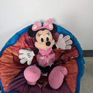 Puede incluir: Un peluche de Minnie Mouse con un vestido rosa con lunares blancos y un lazo a juego. El peluche est&aacute; sentado en un coj&iacute;n colorido con dibujos y un borde azul. El juguete tiene orejas negras, una nariz negra y guantes blancos.