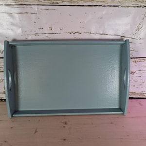 Op de afbeelding: Een rechthoekig, lichtblauw houten dienblad met afgeronde handgrepen. De lade heeft een vlak oppervlak en een licht verhoogde rand. De lade is geschilderd in een effen, matte afwerking.