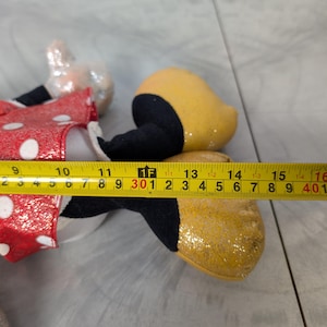 May include: A Minnie Mouse plush toy, featuring a red dress with white polka dots, black shoes, and a yellow glittery shoe. The plush toy measures approximately 15 inches in length.