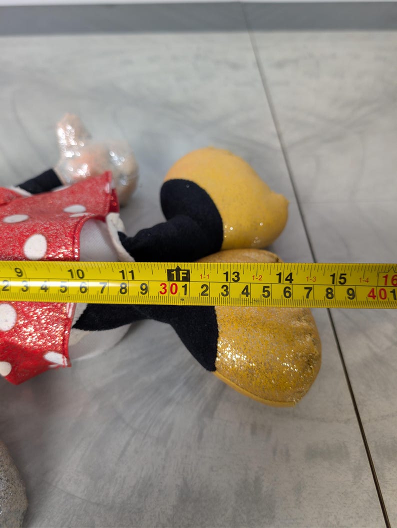 May include: A Minnie Mouse plush toy, featuring a red dress with white polka dots, black shoes, and a yellow glittery shoe. The plush toy measures approximately 15 inches in length.