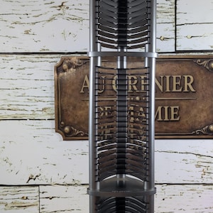 May include: A tall, silver-grey CD tower with a capacity for multiple discs. The tower is made of metal and plastic, with curved slots to hold the CDs. The base is round and sturdy. The background features a sign that reads "AU GRENIER DE LA MIE".