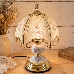 Peut inclure: Une lampe de table de style vintage avec un motif floral. La lampe présente une base et des accents dorés, un centre en céramique blanc et à motifs floraux, et un abat-jour en verre avec un motif floral. La lampe est branchée et allumée, dégageant une lueur chaleureuse.