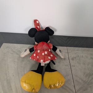 May include: A plush Minnie Mouse doll, viewed from the back, features a red dress with white polka dots and a matching bow. The doll has black ears, yellow glittery shoes, and white gloves with black details. The doll is on a gray surface.