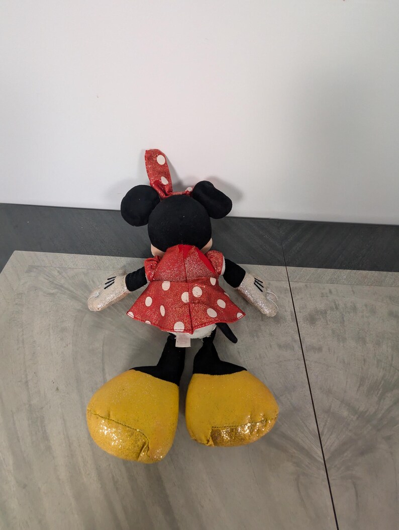 May include: A plush Minnie Mouse doll, viewed from the back, features a red dress with white polka dots and a matching bow. The doll has black ears, yellow glittery shoes, and white gloves with black details. The doll is on a gray surface.
