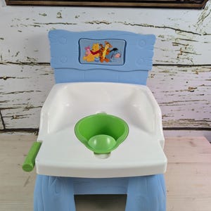 May include: A blue and white plastic potty chair with a Winnie the Pooh design. The chair has a green potty insert and a small handle on the side. The backrest features a rectangular image of Winnie the Pooh and friends.