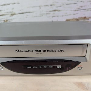 May include: Silver VCR player with the text "DA4 HEAD Hi-Fi VCR 19-MICRON HEADS" and "VHR-H531". The player has a black panel with buttons for power, tape in, VCR, record, timer, and VHS. The VCR also has channel and rewind buttons.