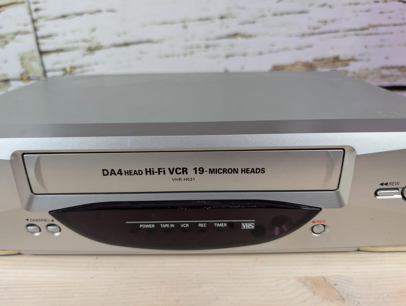 May include: Silver VCR player with the text "DA4 HEAD Hi-Fi VCR 19-MICRON HEADS" and "VHR-H531". The player has a black panel with buttons for power, tape in, VCR, record, timer, and VHS. The VCR also has channel and rewind buttons.