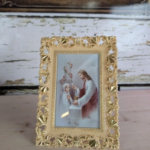 May include: A gold-colored, ornate picture frame with a religious image. The image depicts Jesus Christ offering communion to a young girl. The frame has a detailed, decorative design.