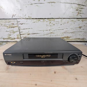 May include: A grey Sharp VCR player with the text "4-HEAD/MID-DRIVE" and "VHS HQ" on the front panel. The VCR has a power button, a power indicator, and a set-up button. The VCR is on a wooden surface.