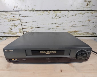 May include: A gray Sharp VCR player with the text "4-HEAD/MID-DRIVE" and "VHS HQ" on the front panel. The VCR has a power button, a power indicator, and a set-up button. The VCR is on a wooden surface.
