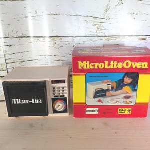 Puede incluir: Un juguete Micro-Lite Oven vintage. El microondas beige y negro tiene una pantalla digital y un dial. La caja roja lleva la inscripción "MicroLite Oven" y muestra a un niño usando el juguete. Para hornear pequeñas comidas y pasteles.