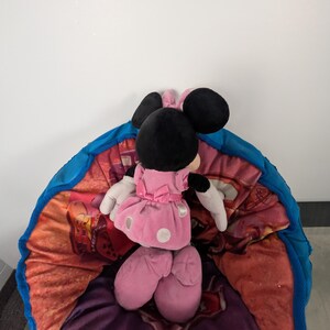Puede incluir: Un peluche de Minnie Mouse con un vestido rosa con lunares blancos, orejas negras y guantes blancos, descansando sobre un puf colorido con borde azul. El puf tiene un dise&ntilde;o floral y de dibujos animados.
