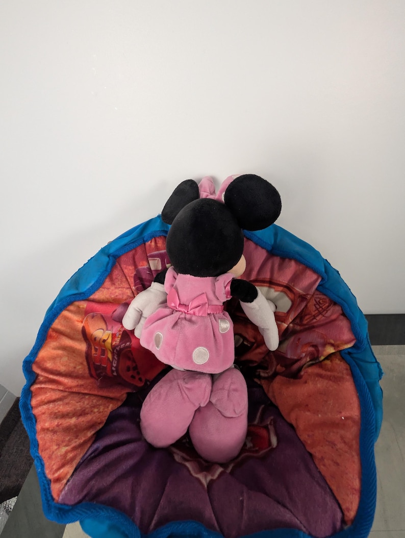 Puede incluir: Un peluche de Minnie Mouse con un vestido rosa con lunares blancos, orejas negras y guantes blancos, descansando sobre un puf colorido con borde azul. El puf tiene un dise&ntilde;o floral y de dibujos animados.