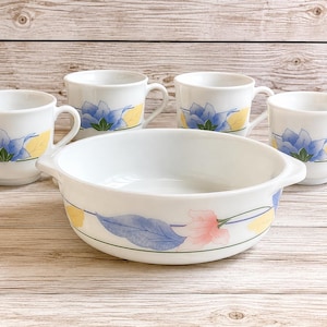 May include: A white ceramic serving bowl with two side handles and four matching mugs, all decorated with a floral pattern featuring blue and pink petals, green leaves, and yellow accents arranged along a thin horizontal line around the exterior.