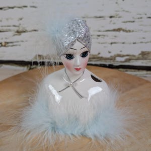 May include: A ceramic art deco-style bust with a silver sequined head covering and a light blue feather accent. The bust features a white body with light blue feather trim and a delicate pearl necklace.