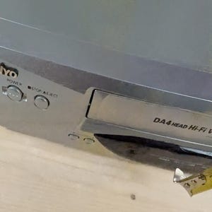 May include: A silver-colored VCR player with the brand name "YO" visible. The player has buttons labeled "POWER" and "STOP/EJECT". The text "DA4 HEAD Hi-Fi VCR" is also visible. A yellow measuring tape is partially in view.