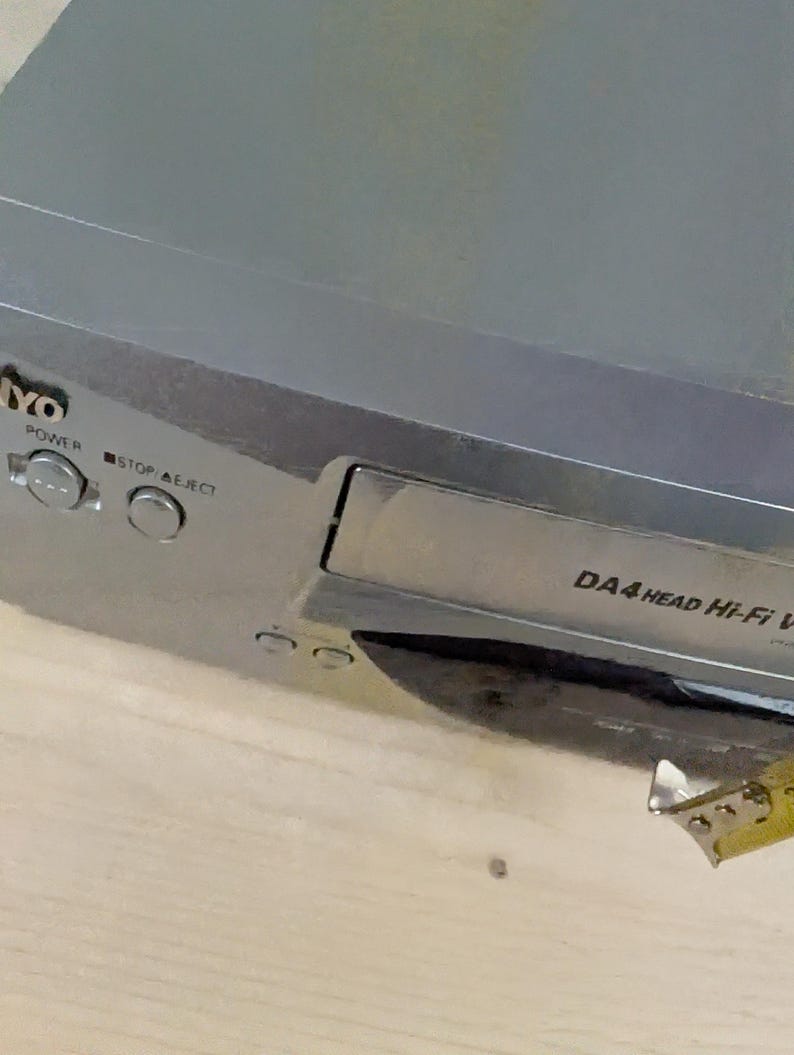 May include: A silver-colored VCR player with the brand name "YO" visible. The player has buttons labeled "POWER" and "STOP/EJECT". The text "DA4 HEAD Hi-Fi VCR" is also visible. A yellow measuring tape is partially in view.