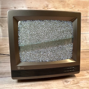 Puede incluir: Un televisor GE gris vintage con una pantalla que muestra estática. El televisor tiene forma rectangular con un marco gris oscuro y una fila de botones en la parte inferior. La palabra "REMOTE" es visible.
