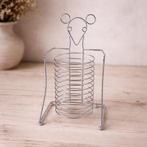 Puede incluir: Un soporte para utensilios de metal plateado con forma de ratón. El soporte tiene un cuerpo cilíndrico con múltiples niveles circulares, brazos y una cabeza con dos orejas circulares. El soporte está diseñado para colocarse de pie sobre una superficie plana.