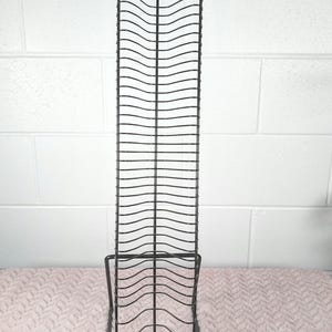 以下が含まれることがあります： 波型のデザインが施された、背の高い黒い金属製のCDラック。ラックは長方形のベースと、CDを保持する一連の湾曲したワイヤーを備えています。ラックは、白いレンガの壁を背景に、ピンク色の表面に立っています。