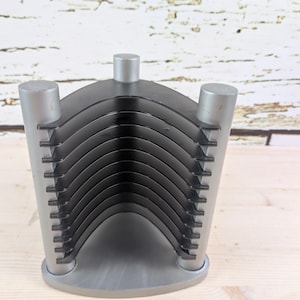 May include: A silver-colored CD holder with a curved design. The holder has a base and three cylindrical supports. The CD holder has a series of black, curved dividers to hold CDs. The background is a light-colored wooden surface.