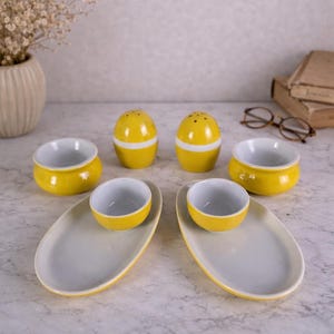 Peut inclure: Un ensemble de plats de service en céramique jaune et blanc. L'ensemble comprend deux salières, deux petits bols, deux grands bols et deux plateaux de service ovales. Les bols et les plateaux ont un intérieur blanc.
