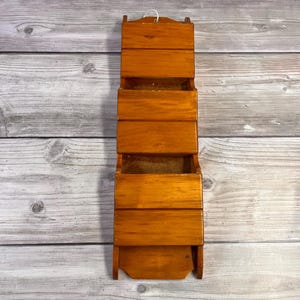May include: A wooden wall-mounted organiser with three compartments and a small shelf at the bottom. The organiser is made of stained wood with a warm, reddish-brown hue. It has a simple, rectangular design with a small loop at the top for hanging.