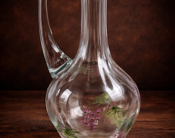 May include: Clear glass pitcher with a curved handle and a pouring spout. The pitcher features a painted design of purple grapes and green leaves. The pitcher is sitting on a wooden surface.